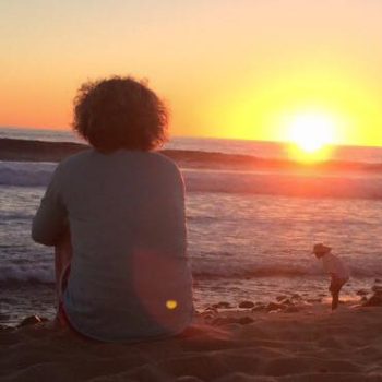 A woman seated on the beach, gazing at the vibrant sunset over the horizon, reflecting tranquility and beauty.