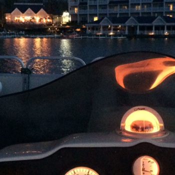 A nighttime view of a boat's dashboard, illuminated instruments and controls against a dark, serene water backdrop.