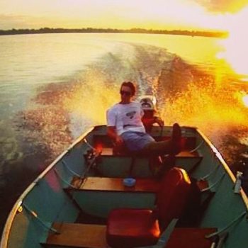 A man in a boat silhouetted against a vibrant sunset, casting warm hues across the tranquil water.